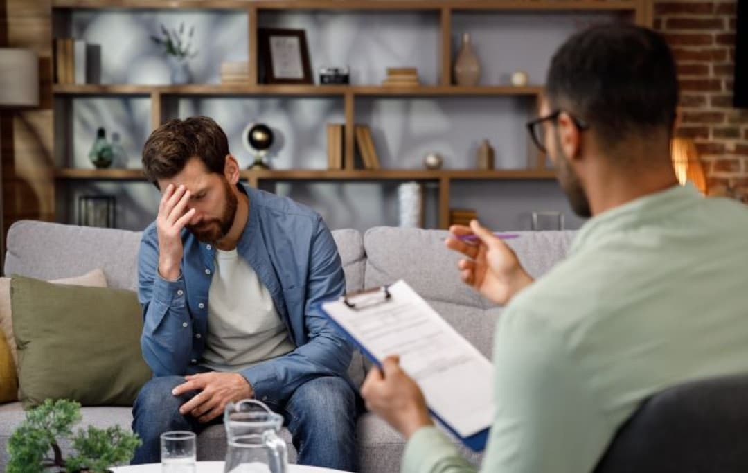 Ein Mann sitzt während einer Therapie auf einer Couch, stützt seinen Kopf mit einer Hand und hat die Augen geschlossen